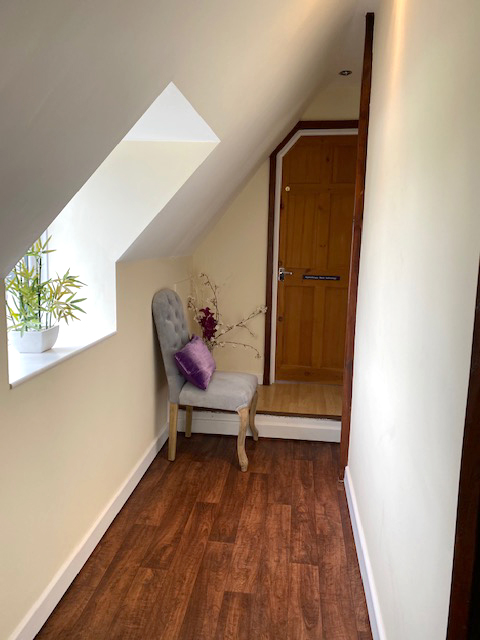 Image of the reception area at Dermedicare aesthetics and skin clinic in Swanland East Yorkshire. Light walls , wooden door, wooden flooring with relaxing light from window to the left  with grey seat and  purple cushion. The scene is adored with lovely flowers.